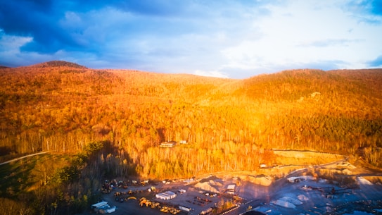 A scenic view of a landscape bathed in warm, golden light. The hillside is covered with dense tree foliage in shades of orange and brown. In the foreground, there is a construction or industrial site with various buildings, vehicles, and materials visible. The sky features a mix of deep blue and soft clouds.
