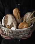 A wicker basket filled with a variety of freshly baked bread, including loaves and rolls, accompanied by wheat stalks and eggs. The basket is being held by hands against a dark background.
