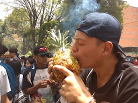 A young man wearing a backward cap is holding and using a pineapple fashioned into a smoking pipe. Smoke is visibly emerging from the top. He is surrounded by a group of people in an outdoor, park-like setting with trees and greenery in the background. The crowd appears diverse and relaxed, with some people engaged in conversation.