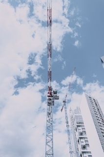 Several construction cranes tower over a partially completed high-rise building. The sky is bright with scattered white clouds, and the metal structure of the cranes stands out against the blue backdrop. The building features modern architecture with glass and steel elements.