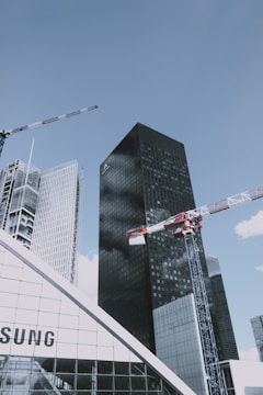 Several skyscrapers tower over the scene, with a clear blue sky as the backdrop. Two large construction cranes intersect in the foreground, indicating ongoing development. The buildings feature modern architecture with sleek glass facades. A prominent billboard or sign partially visible at the bottom left adds to the urban atmosphere.