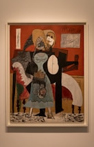 A painting featuring abstract human figures with geometric forms and distorted features. The background includes a red wall, light-colored frame, and elements suggestive of furniture. The figures are rendered in contrasting shades, with black, white, and various earthy tones. The style is reminiscent of cubism, characterized by fragmented shapes and overlapping planes.