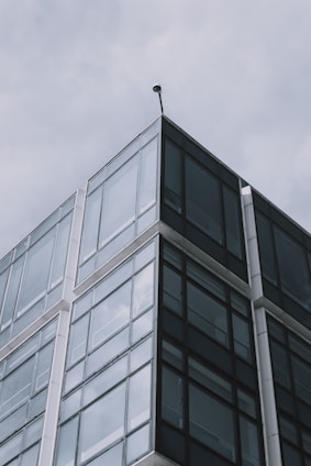 A modern building features a sharp corner with large glass windows extending upward against a cloudy sky backdrop. The structure has a sleek, reflective surface with metallic framing, emphasizing contemporary architectural design.