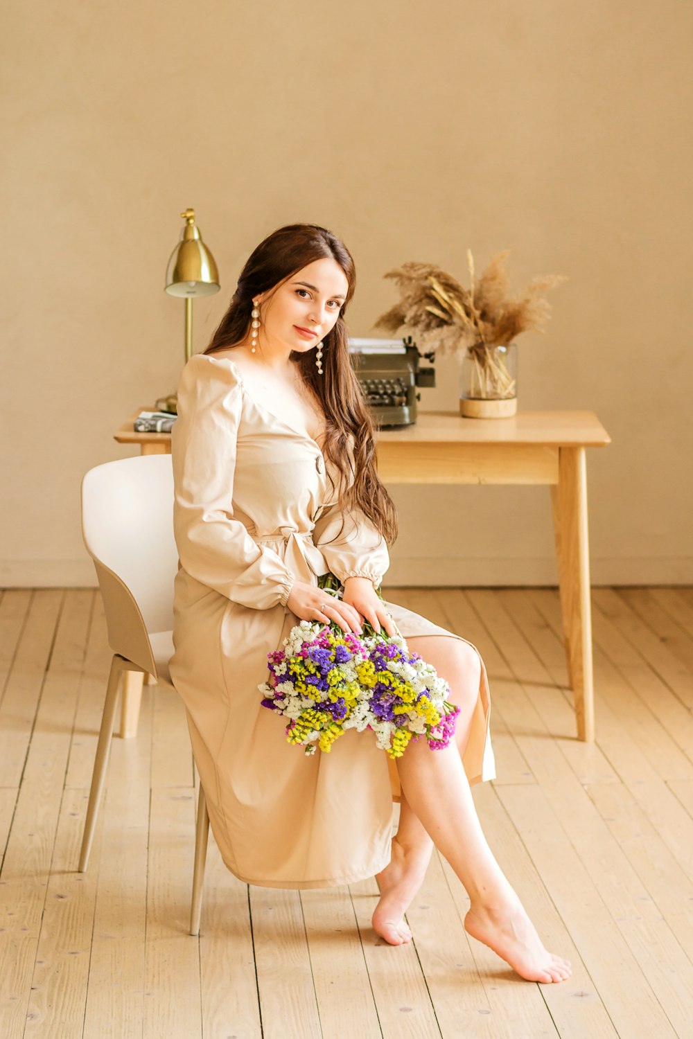 Mujer descalza con vestido blanco de manga larga sentada en una silla cerca de una mesa rectangular de madera beige