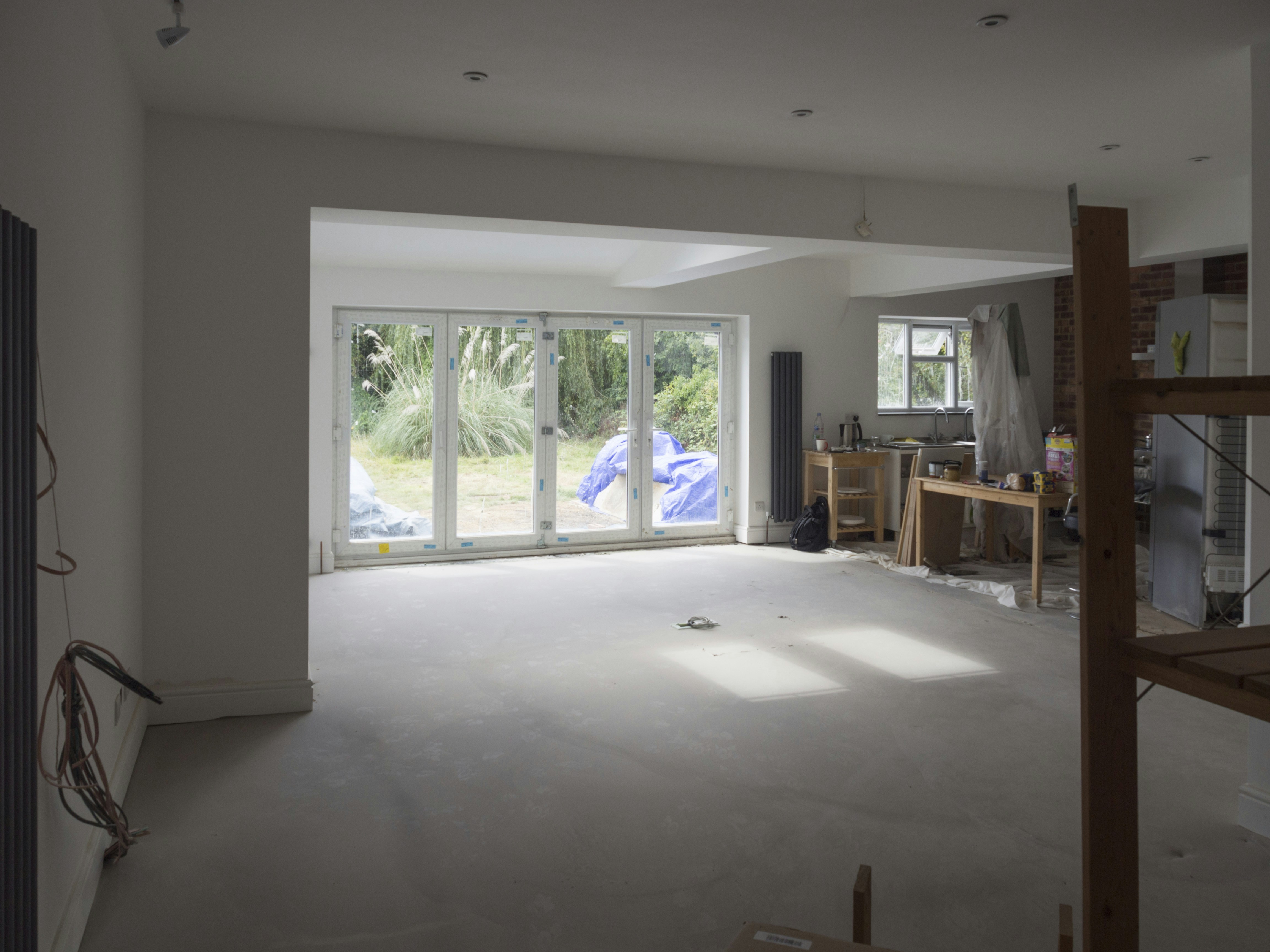 A partially renovated room with large windows overlooking a garden. The room is mostly empty, with some construction materials like boards and plastic wrapped furniture on the right side. There are cables hanging from the ceiling and a small workbench with tools near the window on the right.
