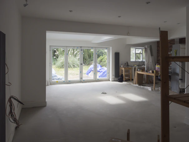 A partially renovated room with large windows overlooking a garden. The room is mostly empty, with some construction materials like boards and plastic wrapped furniture on the right side. There are cables hanging from the ceiling and a small workbench with tools near the window on the right.