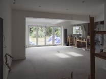 A partially renovated room with large windows overlooking a garden. The room is mostly empty, with some construction materials like boards and plastic wrapped furniture on the right side. There are cables hanging from the ceiling and a small workbench with tools near the window on the right.