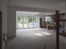 A partially renovated room with large windows overlooking a garden. The room is mostly empty, with some construction materials like boards and plastic wrapped furniture on the right side. There are cables hanging from the ceiling and a small workbench with tools near the window on the right.