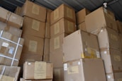 A large stack of brown cardboard boxes piled up in a storage area. The boxes are sealed with tape and have various labels and shipping details attached. The ceiling and a small surveillance camera are visible above the stacked boxes.