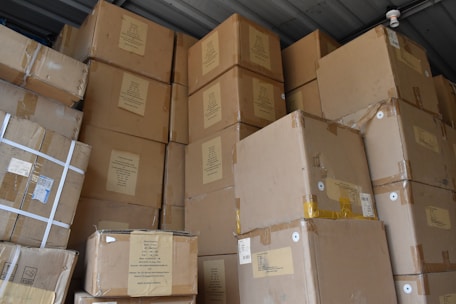 A large stack of brown cardboard boxes piled up in a storage area. The boxes are sealed with tape and have various labels and shipping details attached. The ceiling and a small surveillance camera are visible above the stacked boxes.