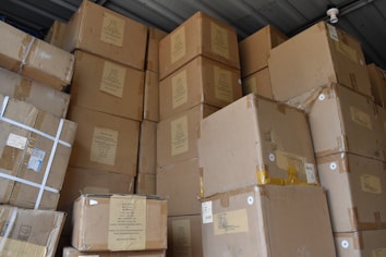 A large stack of brown cardboard boxes piled up in a storage area. The boxes are sealed with tape and have various labels and shipping details attached. The ceiling and a small surveillance camera are visible above the stacked boxes.