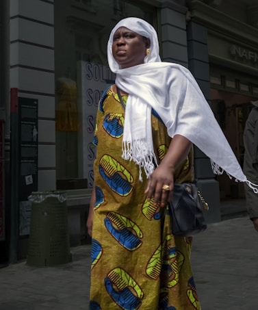 A person wearing a colorful traditional garment with bold patterns and a white headscarf. They are holding a black bag. The background includes a partially visible storefront with signage and a green trash can.