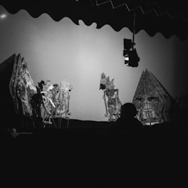 Intricately designed shadow puppets are displayed against a lit backdrop creating an artistic scene. The silhouettes of the puppets showcase detailed patterns and elaborate costumes, characteristic of a traditional shadow play. The lighting is set to emphasize the contrast between the puppets and the screen behind them, with a dark figure sitting among the audience.
