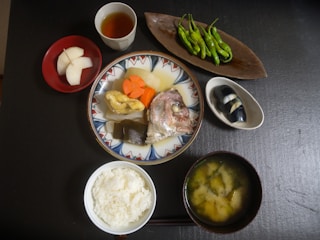 A traditional Japanese meal displayed on a dark table. It includes a central dish with a variety of items, such as a fish head, carrots, tofu, and seaweed. Surrounding it are smaller bowls containing a cup of tea, some pickled vegetables, a bowl of white rice, green peppers on a decorative plate, and a bowl of miso soup.