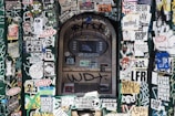 An urban scene featuring an ATM covered in layers of stickers and graffiti. The surrounding area is densely packed with colorful and monochrome stickers displaying various logos, illustrations, and text. The wall and ATM surfaces are heavily marked with graffiti, adding to the chaotic and vibrant aesthetic.
