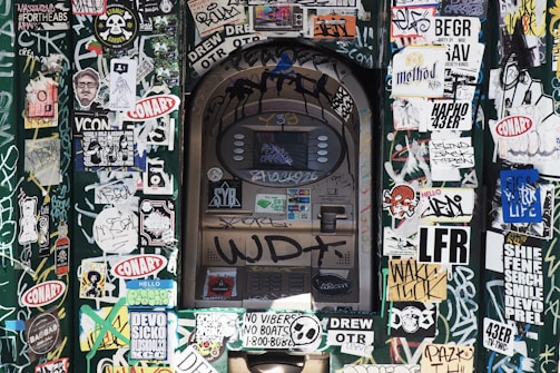 An urban scene featuring an ATM covered in layers of stickers and graffiti. The surrounding area is densely packed with colorful and monochrome stickers displaying various logos, illustrations, and text. The wall and ATM surfaces are heavily marked with graffiti, adding to the chaotic and vibrant aesthetic.