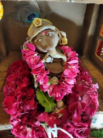 A small handcrafted clay figure decorated with vibrant pink flower garlands and intricate jewelry. The figure has a prominent trunk-like feature, a gold headpiece adorned with a feather, and small sparkling embellishments. The clay is earthy brown, contrasting with the bright florals surrounding it.