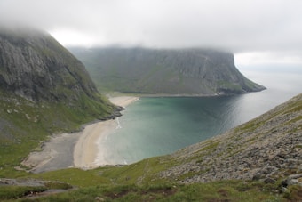 A scenic view of a beach nestled between grassy mountains with a cloudy sky. The water is a clear turquoise, fading into a deeper blue as it stretches toward the horizon. Stone cliffs rise sharply from the sandy beach, partially obscured by low-hanging clouds. The landscape is lush and rugged, evoking a sense of solitude and natural beauty.