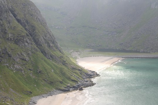A serene landscape featuring a coastline with turquoise water gently meeting a sandy beach. Rugged cliffs covered in green vegetation rise sharply next to the beach, and the scene is enveloped in a soft light, enhancing its tranquility.