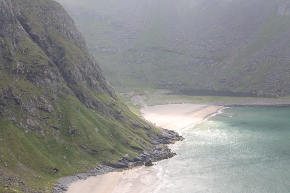 A serene landscape featuring a coastline with turquoise water gently meeting a sandy beach. Rugged cliffs covered in green vegetation rise sharply next to the beach, and the scene is enveloped in a soft light, enhancing its tranquility.