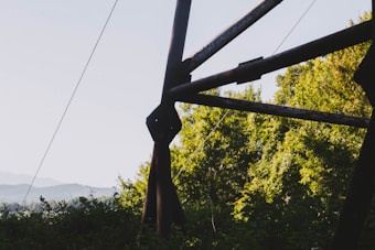 Rustic metal beams intersect against a backdrop of vibrant green trees, suggesting an outdoor industrial or construction setting. Soft hills are visible in the distant background under a partly cloudy sky.