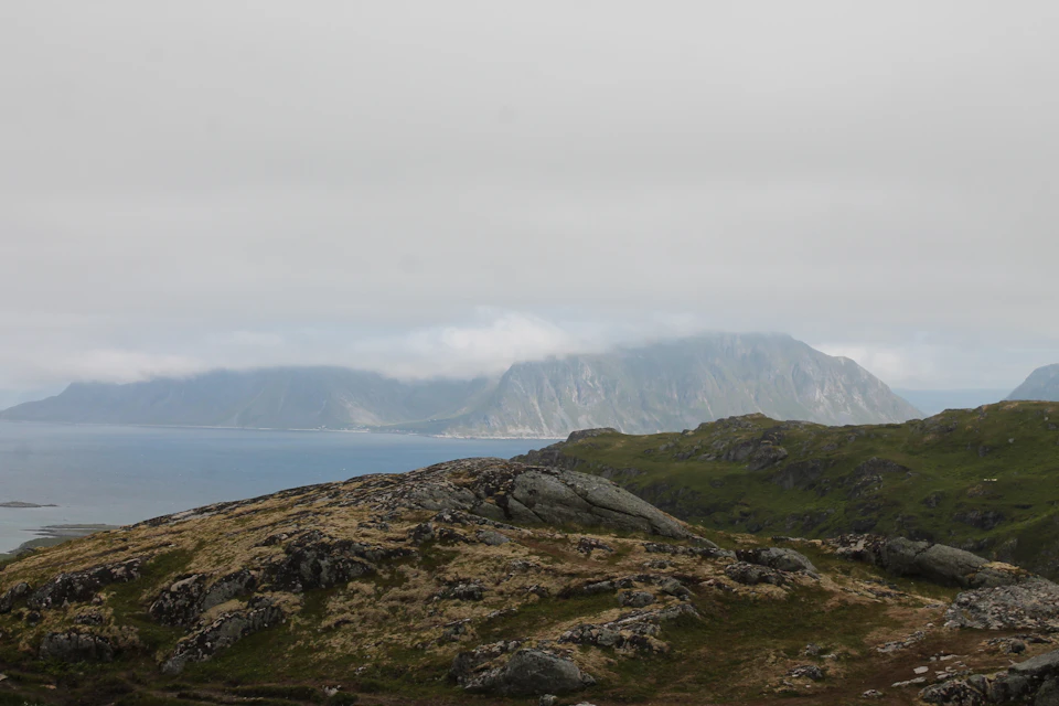 The image depicts a serene landscape with a rocky hillside covered in lush green grass, overlooking a vast body of water. The sky above is overcast, casting a soft light on the scene and creating an ethereal atmosphere. In the distance, several islands can be seen, adding depth to the image.