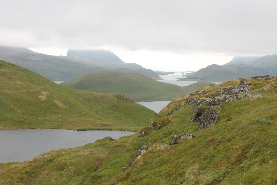 The image depicts a serene landscape with a large lake nestled amidst rolling hills and mountains. The lake is surrounded by lush green grass, creating a tranquil atmosphere. In the distance, several rocky outcrops can be seen, adding to the rugged beauty of the scene. The sky above is overcast, casting a soft light on the landscape below.