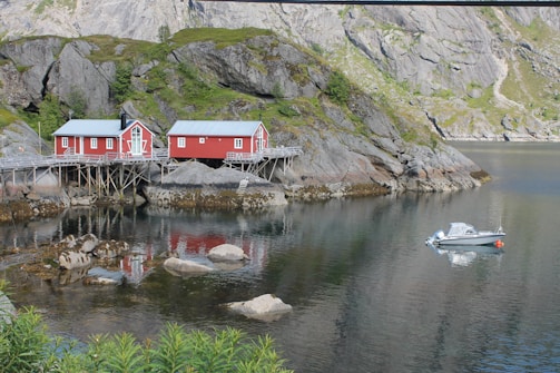 Two red wooden cabins are built on stilts along the rocky shoreline of a calm body of water. The surrounding area is characterized by steep, rocky hills with patches of green vegetation. A small motorboat is moored nearby, floating on the reflective water surface.