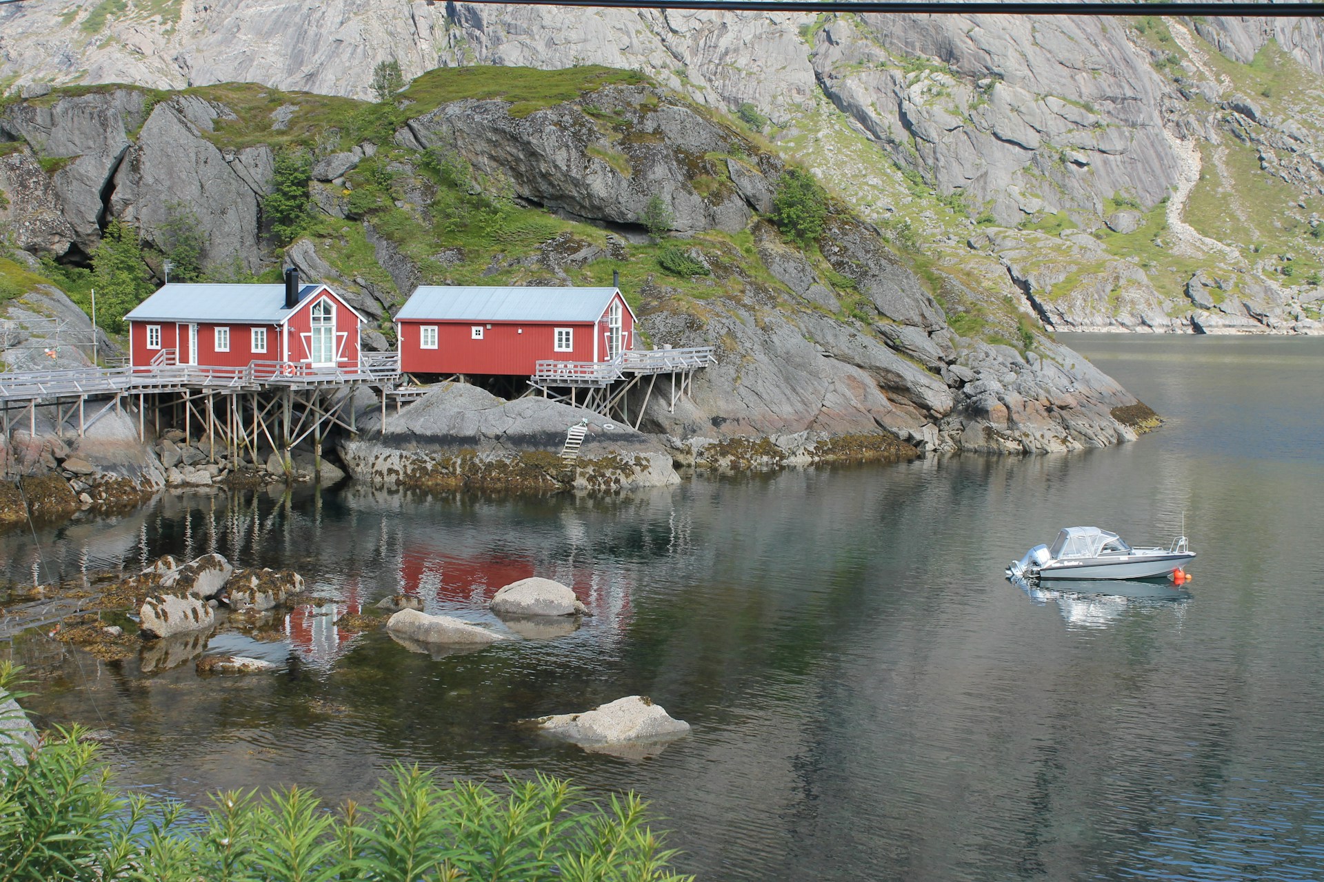 Two red wooden cabins are built on stilts along the rocky shoreline of a calm body of water. The surrounding area is characterized by steep, rocky hills with patches of green vegetation. A small motorboat is moored nearby, floating on the reflective water surface.