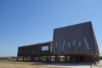 A modern building with a unique architectural design, featuring a large, angular wooden structure elevated on stilts. The building's facade includes several narrow, vertical windows. The surrounding landscape consists of dry grass and open skies.