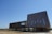 A modern building with a unique architectural design, featuring a large, angular wooden structure elevated on stilts. The building's facade includes several narrow, vertical windows. The surrounding landscape consists of dry grass and open skies.