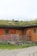 Several sheep graze on a grassy roof of a wooden barn. The barn has a rustic appearance with a natural wood finish and small green-framed windows. There is a metal gate and some wooden crates near the barn.