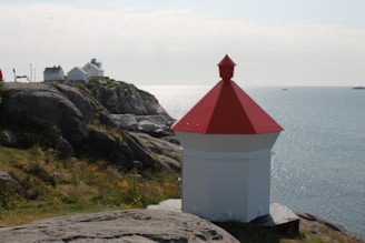 A small red and white lighthouse stands on rocky terrain overlooking the ocean. In the background, there are two white houses perched on a larger rocky promontory, surrounded by a grassy landscape with scattered yellow flowers.