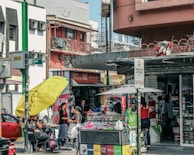 A busy urban street scene with multiple people engaged in various activities. There is a storefront with advertisements and signs in Spanish, and a stand with a large yellow umbrella. Two women appear to be conversing near the stand, while others are shopping or walking by. Vehicles are parked in the background, indicating a city environment.