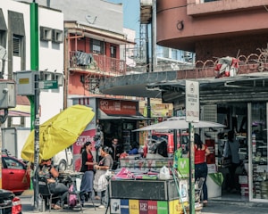A busy urban street scene with multiple people engaged in various activities. There is a storefront with advertisements and signs in Spanish, and a stand with a large yellow umbrella. Two women appear to be conversing near the stand, while others are shopping or walking by. Vehicles are parked in the background, indicating a city environment.