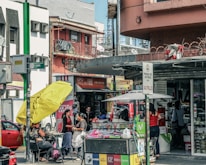 A busy urban street scene with multiple people engaged in various activities. There is a storefront with advertisements and signs in Spanish, and a stand with a large yellow umbrella. Two women appear to be conversing near the stand, while others are shopping or walking by. Vehicles are parked in the background, indicating a city environment.