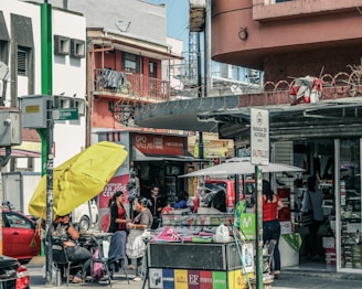 A busy urban street scene with multiple people engaged in various activities. There is a storefront with advertisements and signs in Spanish, and a stand with a large yellow umbrella. Two women appear to be conversing near the stand, while others are shopping or walking by. Vehicles are parked in the background, indicating a city environment.