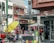 A busy urban street scene with multiple people engaged in various activities. There is a storefront with advertisements and signs in Spanish, and a stand with a large yellow umbrella. Two women appear to be conversing near the stand, while others are shopping or walking by. Vehicles are parked in the background, indicating a city environment.