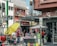 A busy urban street scene with multiple people engaged in various activities. There is a storefront with advertisements and signs in Spanish, and a stand with a large yellow umbrella. Two women appear to be conversing near the stand, while others are shopping or walking by. Vehicles are parked in the background, indicating a city environment.