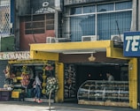 A street scene featuring a small produce stand with an assortment of fruits and vegetables displayed outside. Adjacent is a bakery with a colorful canopy and a glass display case filled with various baked goods. A person stands inside the bakery near the counter, while another person is outside near the produce. The exterior of the building shows signs of wear, with air conditioning units on top.