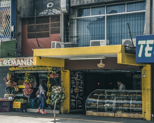 A street scene featuring a small produce stand with an assortment of fruits and vegetables displayed outside. Adjacent is a bakery with a colorful canopy and a glass display case filled with various baked goods. A person stands inside the bakery near the counter, while another person is outside near the produce. The exterior of the building shows signs of wear, with air conditioning units on top.