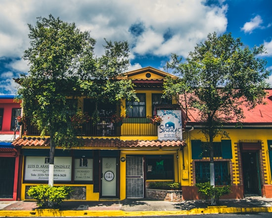A vibrant building with a yellow facade and a red roof, surrounded by leafy trees and blue skies. The structure has a sign for a Shakti establishment and another sign for a lawyer's office. The windows are framed with green shutters, and there are several plants in front of the building.