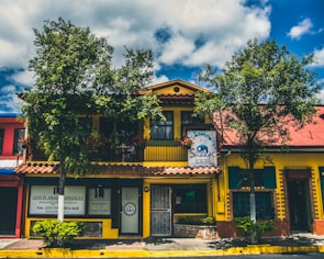 A vibrant building with a yellow facade and a red roof, surrounded by leafy trees and blue skies. The structure has a sign for a Shakti establishment and another sign for a lawyer's office. The windows are framed with green shutters, and there are several plants in front of the building.