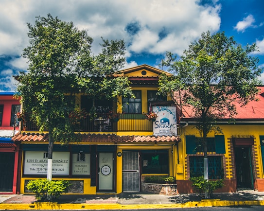 A vibrant building with a yellow facade and a red roof, surrounded by leafy trees and blue skies. The structure has a sign for a Shakti establishment and another sign for a lawyer's office. The windows are framed with green shutters, and there are several plants in front of the building.