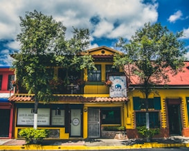 A vibrant building with a yellow facade and a red roof, surrounded by leafy trees and blue skies. The structure has a sign for a Shakti establishment and another sign for a lawyer's office. The windows are framed with green shutters, and there are several plants in front of the building.