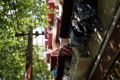 A street view of an urban residential area with clothes hanging to dry on balconies. Red and white awnings are visible above the balconies. The scene is set against lush green trees, and power lines run across the sky.