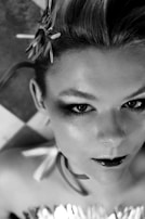 A black and white photograph showcasing a person with styled hair adorned with decorative elements, intense eye makeup, and glossy lips. The background features a geometric tiled pattern.