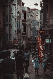 A narrow, bustling street with people walking along sidewalks between tall, closely-packed urban buildings. The architecture features multiple stories with various windows and balconies. Street lights hang overhead, and a large red Vodafone flag stands out in the scene. Several shops have illuminated signs in different languages.