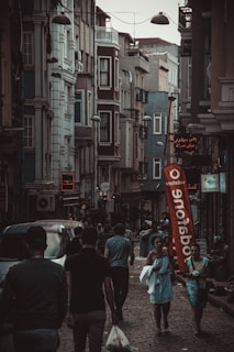 A narrow, bustling street with people walking along sidewalks between tall, closely-packed urban buildings. The architecture features multiple stories with various windows and balconies. Street lights hang overhead, and a large red Vodafone flag stands out in the scene. Several shops have illuminated signs in different languages.