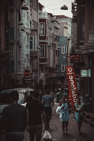 A narrow, bustling street with people walking along sidewalks between tall, closely-packed urban buildings. The architecture features multiple stories with various windows and balconies. Street lights hang overhead, and a large red Vodafone flag stands out in the scene. Several shops have illuminated signs in different languages.